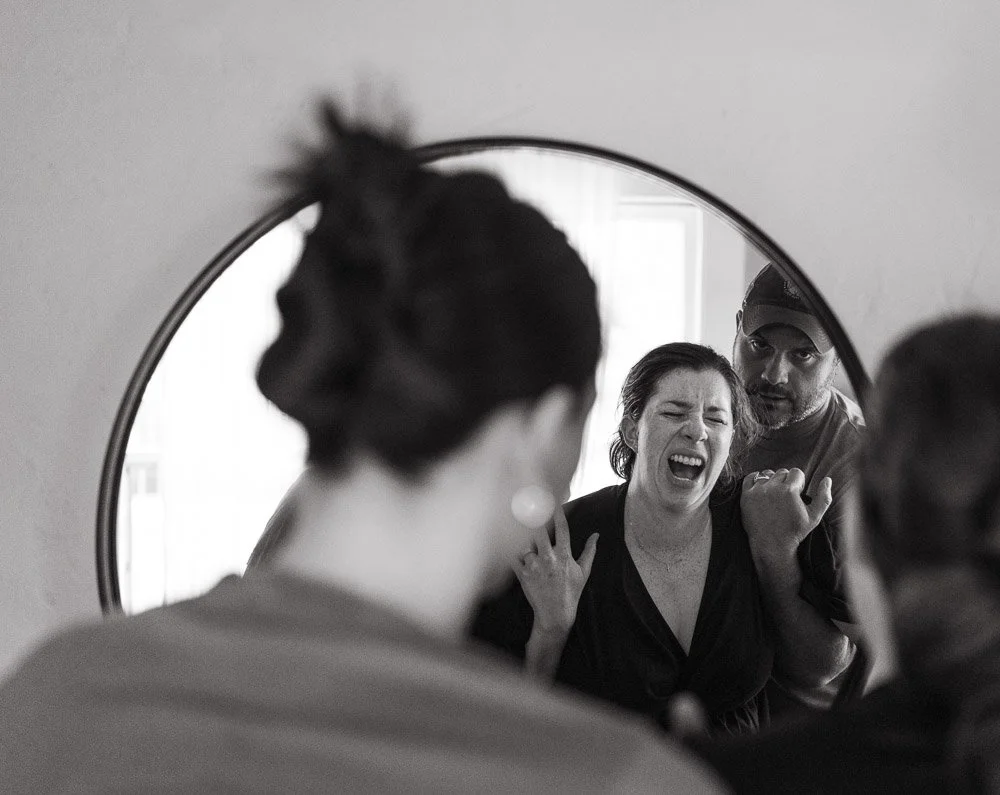 A woman crying and a man with a serious expression look into a mirror, with the woman's reflection showing her crying and the man's face next to her.