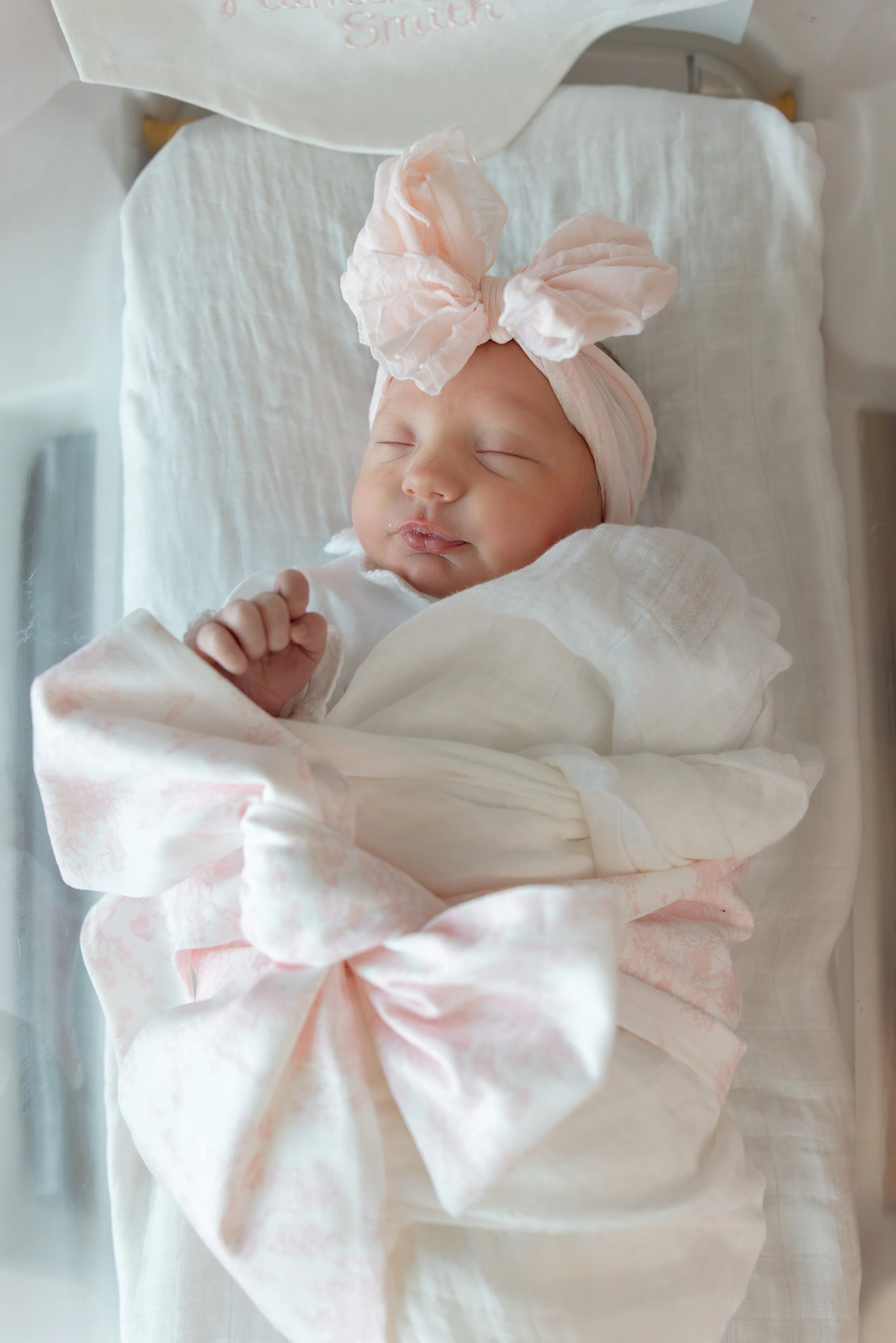 in natural lighting, a newborn baby girl is wearing a pink bow and swaddled in a monogrammed pink and white bow outfit while sleeping in a hospital bassinet. 