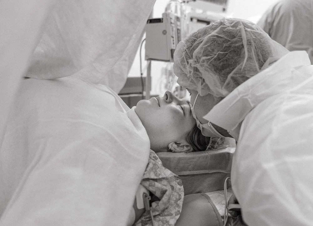 Austin birth photographer documentary image of husband kissing his wife's head while she's laying on the operating table about to have a c-section, black and white image