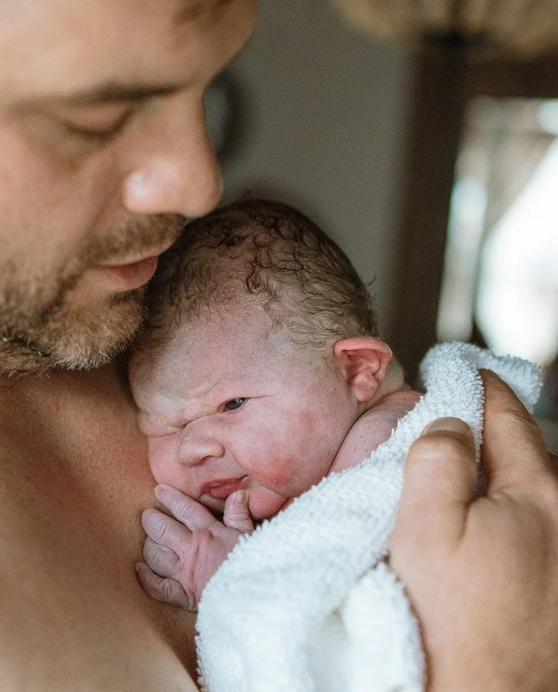 A man holding a newborn baby close to his chest, with the baby's face resting against him. The baby has wet hair and is wrapped in a white towel.