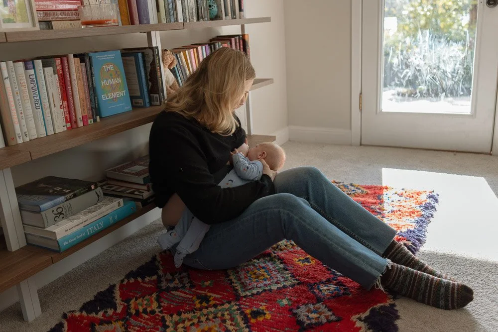 A mom sits on a colorful carpet in here home while leaning against a bookshelf and nursing her newborn baby. 