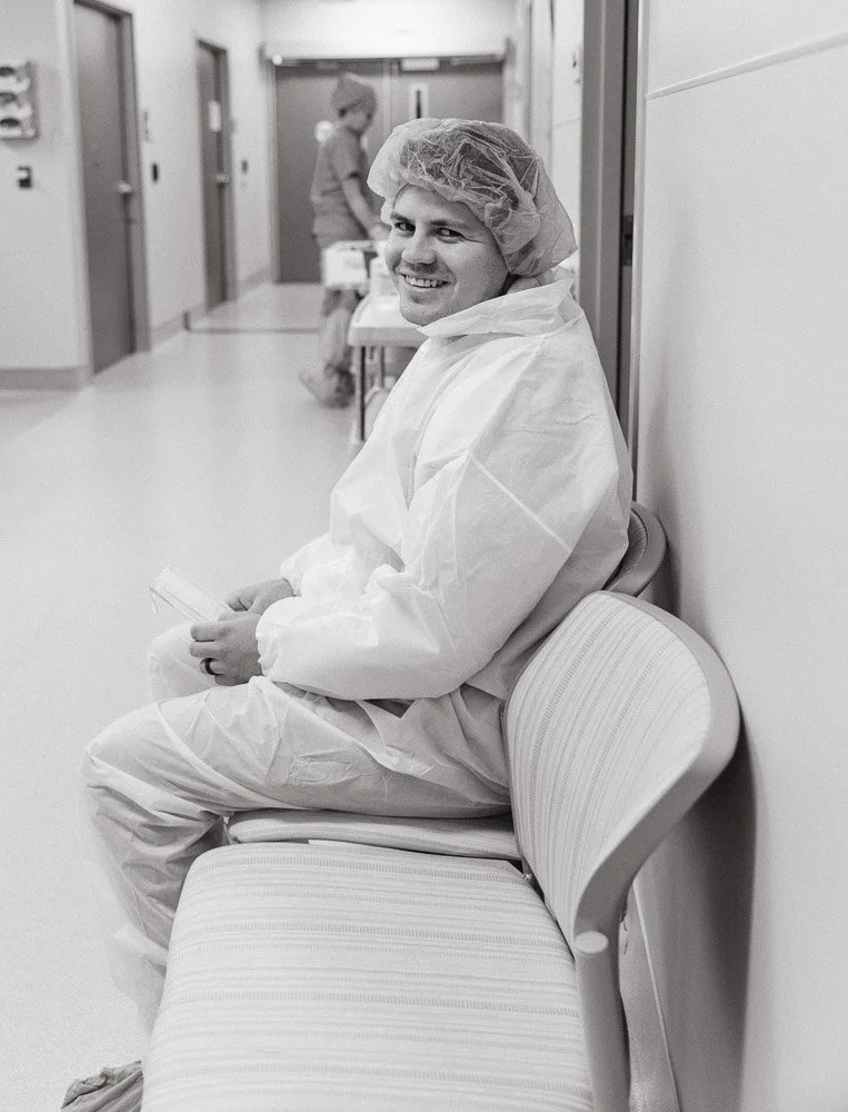 A man in medical scrubs and a hairnet sitting on a hospital waiting area bench, smiling at the camera, with a nurse in the background.