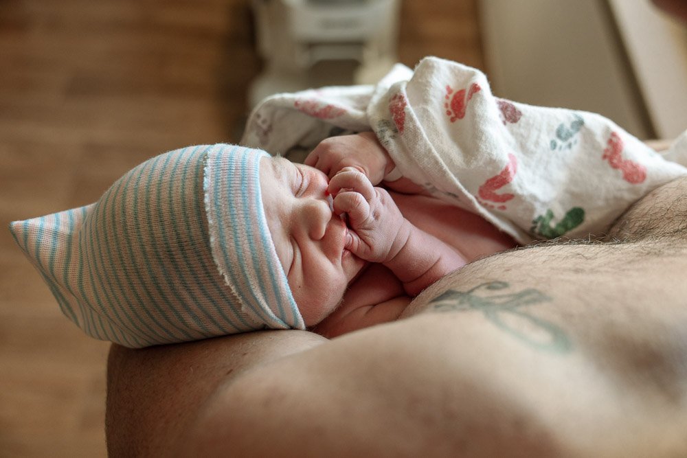 A newborn baby wearing a striped hospital hat, snuggled against an adult, with a hospital blanket partially covering them.