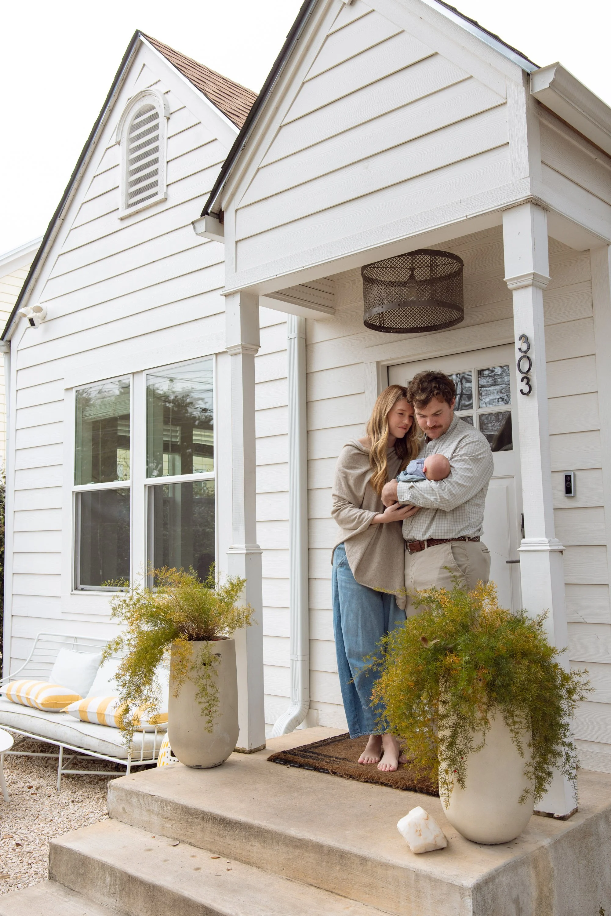 A couple standing on a porch holding a newborn baby outside a white house with potted plants and outdoor furniture.