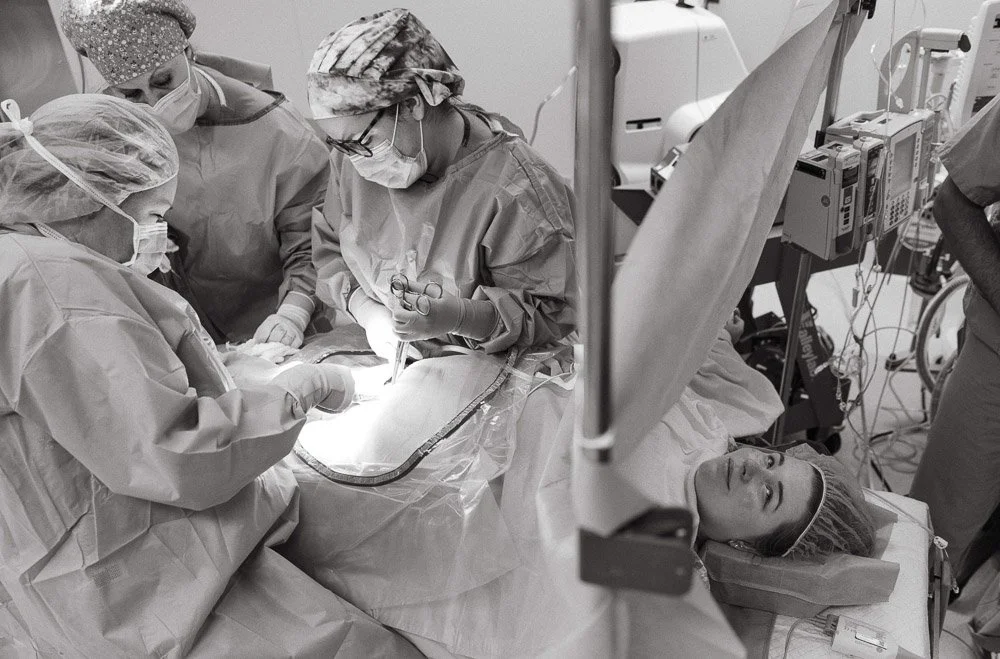 Medical team performing surgery on a patient in an operating room, with a woman lying on the operating table and wearing surgical attire.