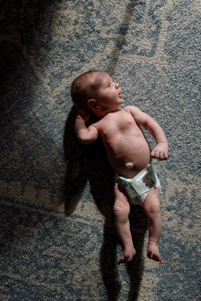 A newborn baby lying on a textured gray and blue rug, looking to the side with a relaxed expression, wearing only a diaper.