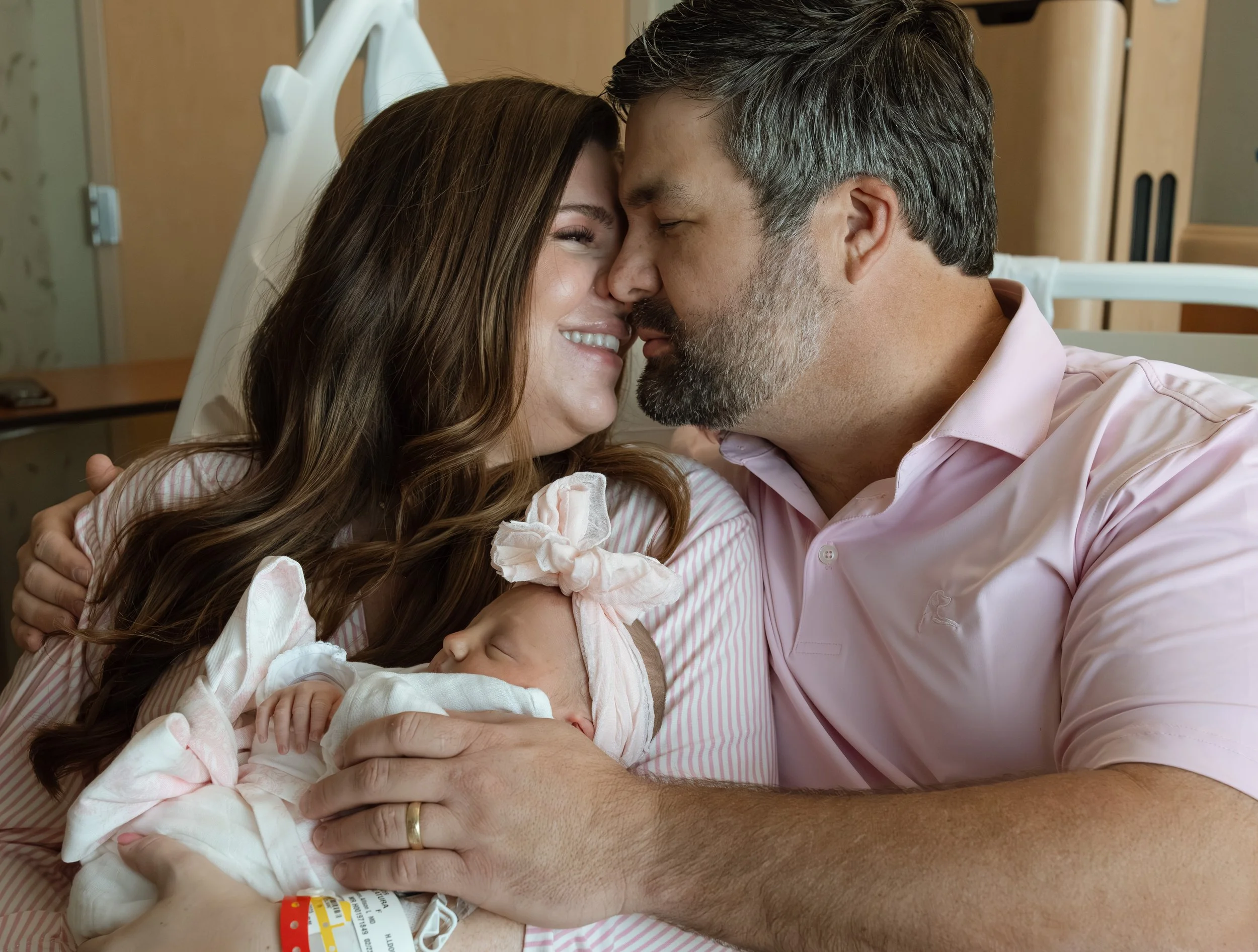 a smiling woman holding a newborn baby girl in a pink bow headband leans in for a kiss from her husband who is wearing a pink shirt, both sitting on a hospital bed. 