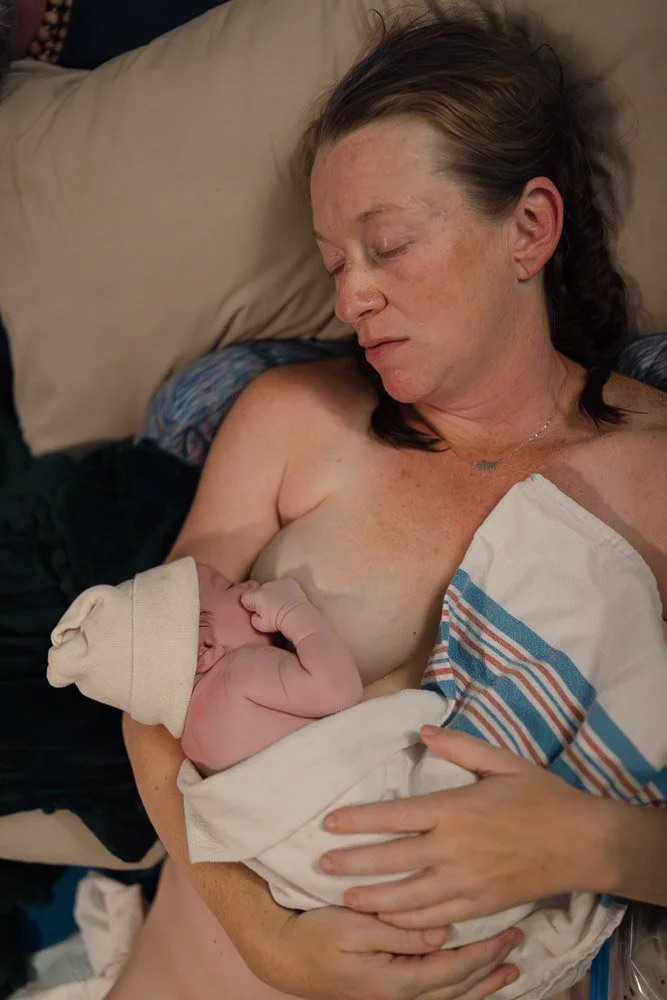 A woman with light skin and dark hair, lying on a pillow, breastfeeding a newborn baby wrapped in a hospital blanket. The woman is topless, with her eyes closed, and appears to be resting.