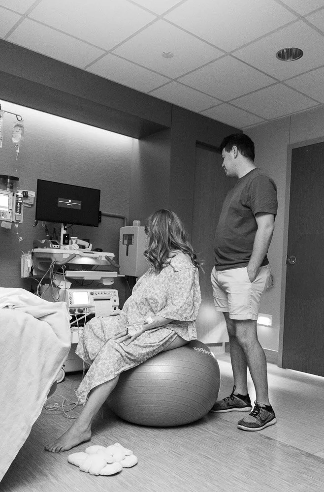 A woman in hospital gown sitting on an exercise ball in a hospital room, with a man in casual clothes standing nearby.