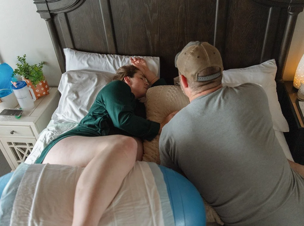 A woman is lying sideways on a bed with her eyes closed, resting her head on a pillow. A man, wearing a gray T-shirt and a beige cap, is sitting beside her with his back to the camera, looking at her. The bed has white pillows, and there is a dark wo