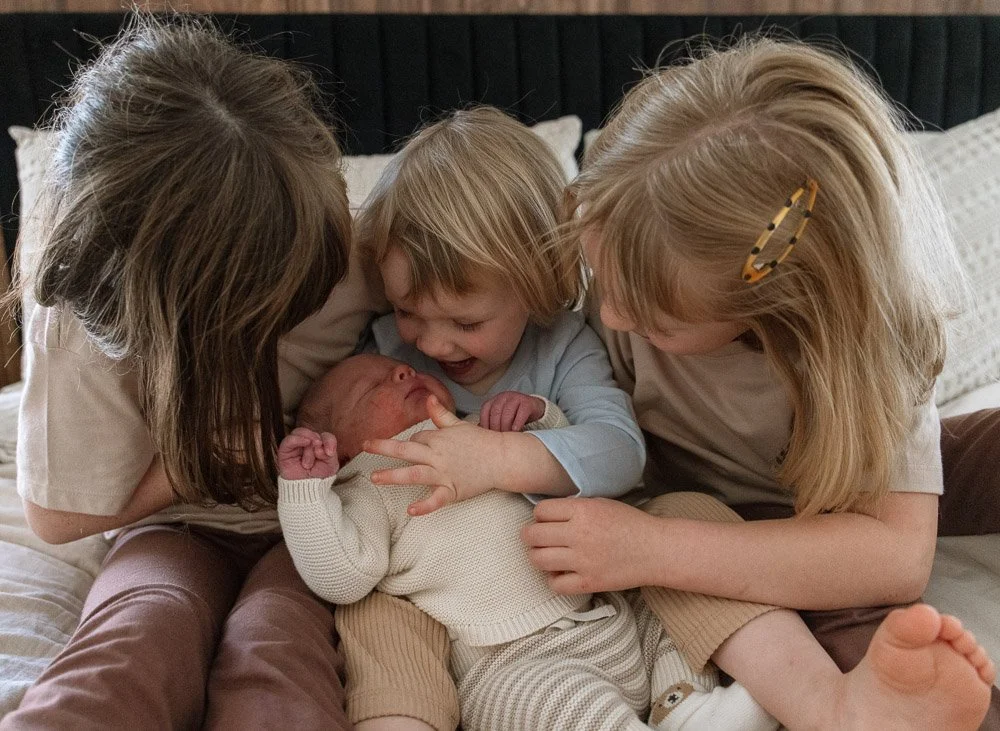 Two young children and an adult girl gathered around a newborn baby on a bed, smiling and touching the baby.