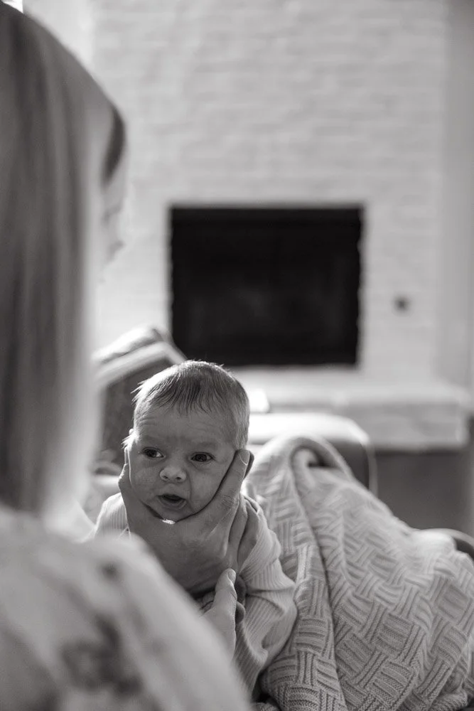 A baby lying on a caregiver's lap, looking at the caregiver, in a cozy room with a fireplace in the background.