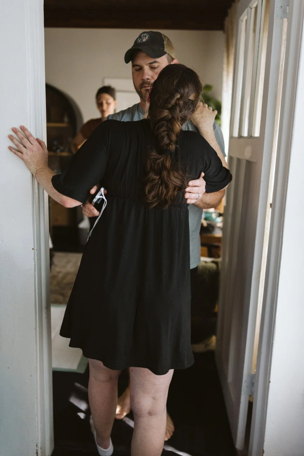A woman with braided hair hugging a man wearing a baseball cap, in a doorway.