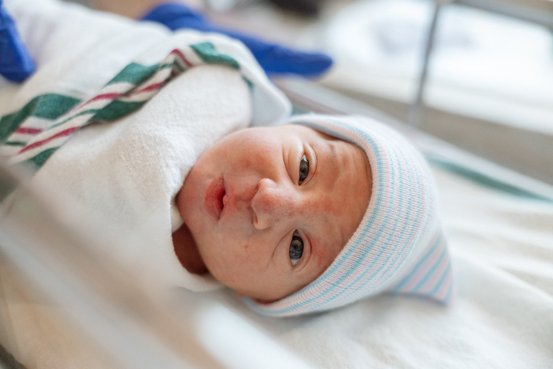 A newborn baby lying in a hospital bed, wearing a striped hospital cap and wrapped in a white hospital blanket.