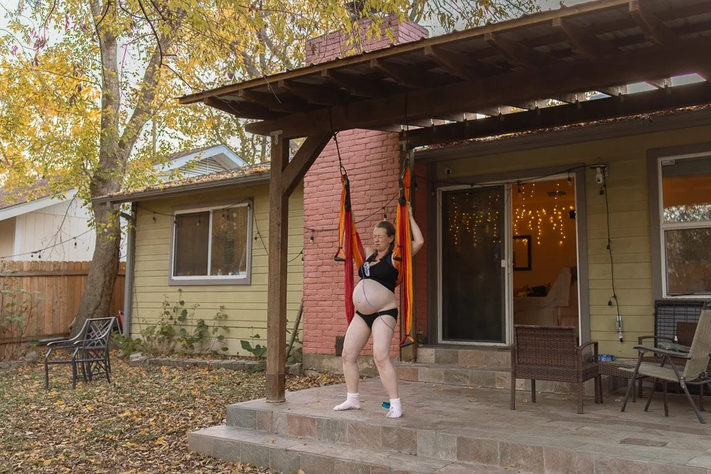A woman on a backyard patio, wearing black lingerie, socks, and a harness, holding onto hammock straps hanging from a wooden pergola, with a house, trees, and string lights visible in the background.