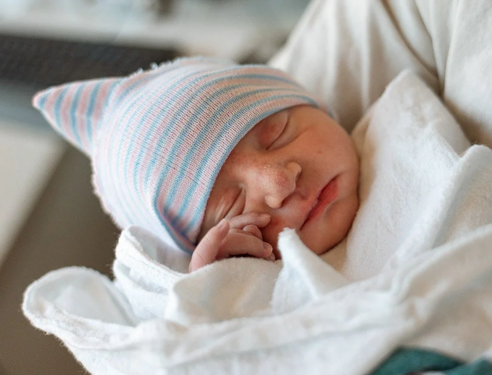 Close-up of a newborn baby sleeping peacefully, wearing a striped pastel beanie, swaddled in white cloth.