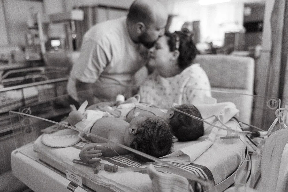 A mother and father holding hands and leaning their foreheads together in a hospital room, with two tiny, premature newborn twins in an incubator in the foreground, the babies are in the NICU