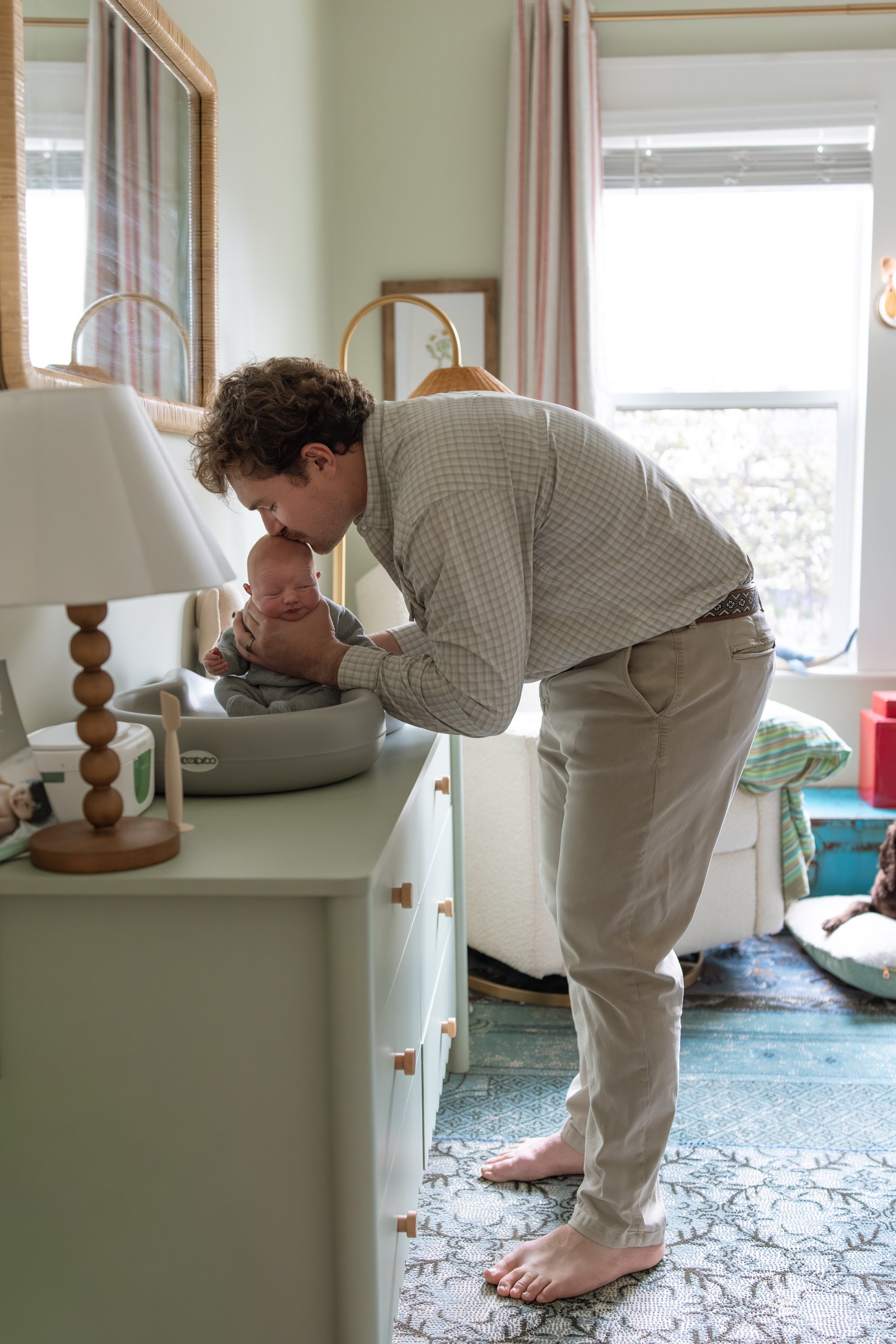 dad and newborn in the nursery with dad leaning over kissing the baby's head- nursery is professionally decorated by an interior designer and has lots of greens and blues. 