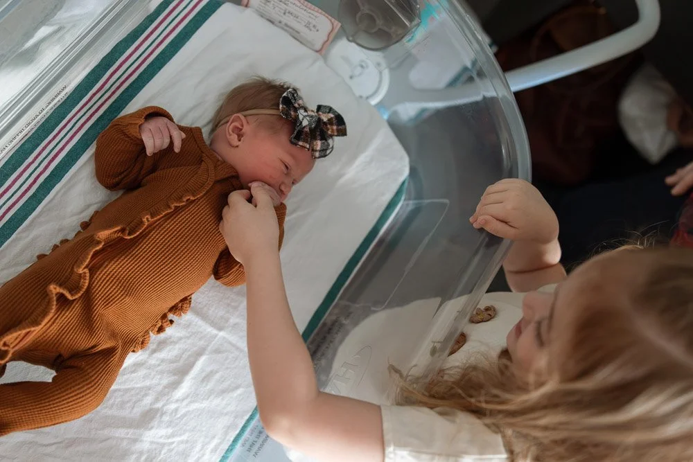 A young girl is reaching into an incubator and gently touching the hand of a newborn baby lying inside.