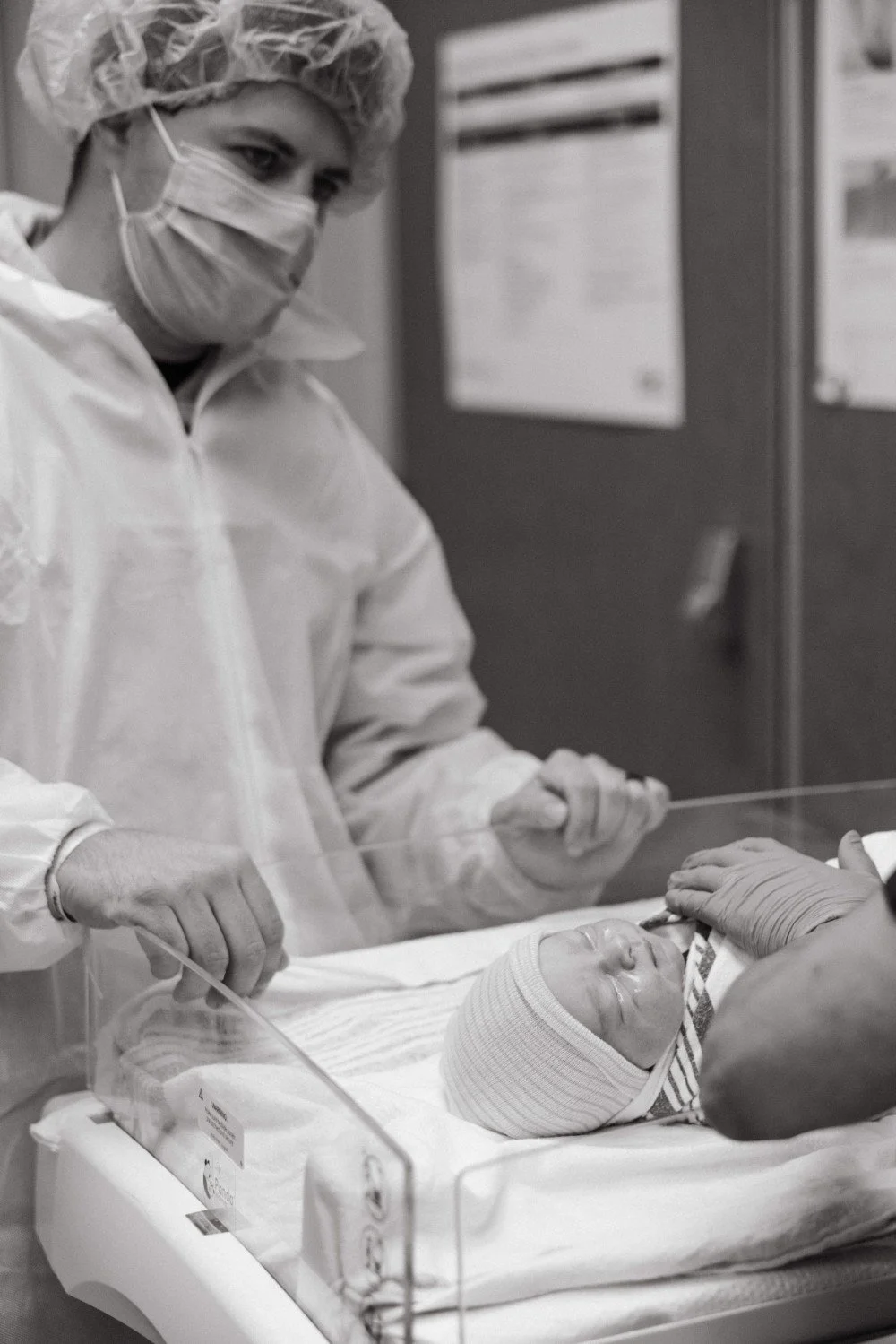 A healthcare worker dressed in protective gear, including a mask, gloves, and a gown, is viewing a newborn baby in a hospital bassinet.