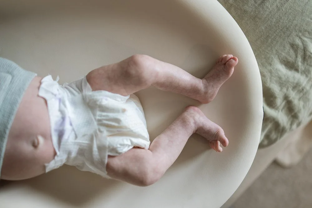 Close-up of a newborn baby's legs and torso lying on a cream-colored surface, wearing a white diaper and a light-colored shirt.