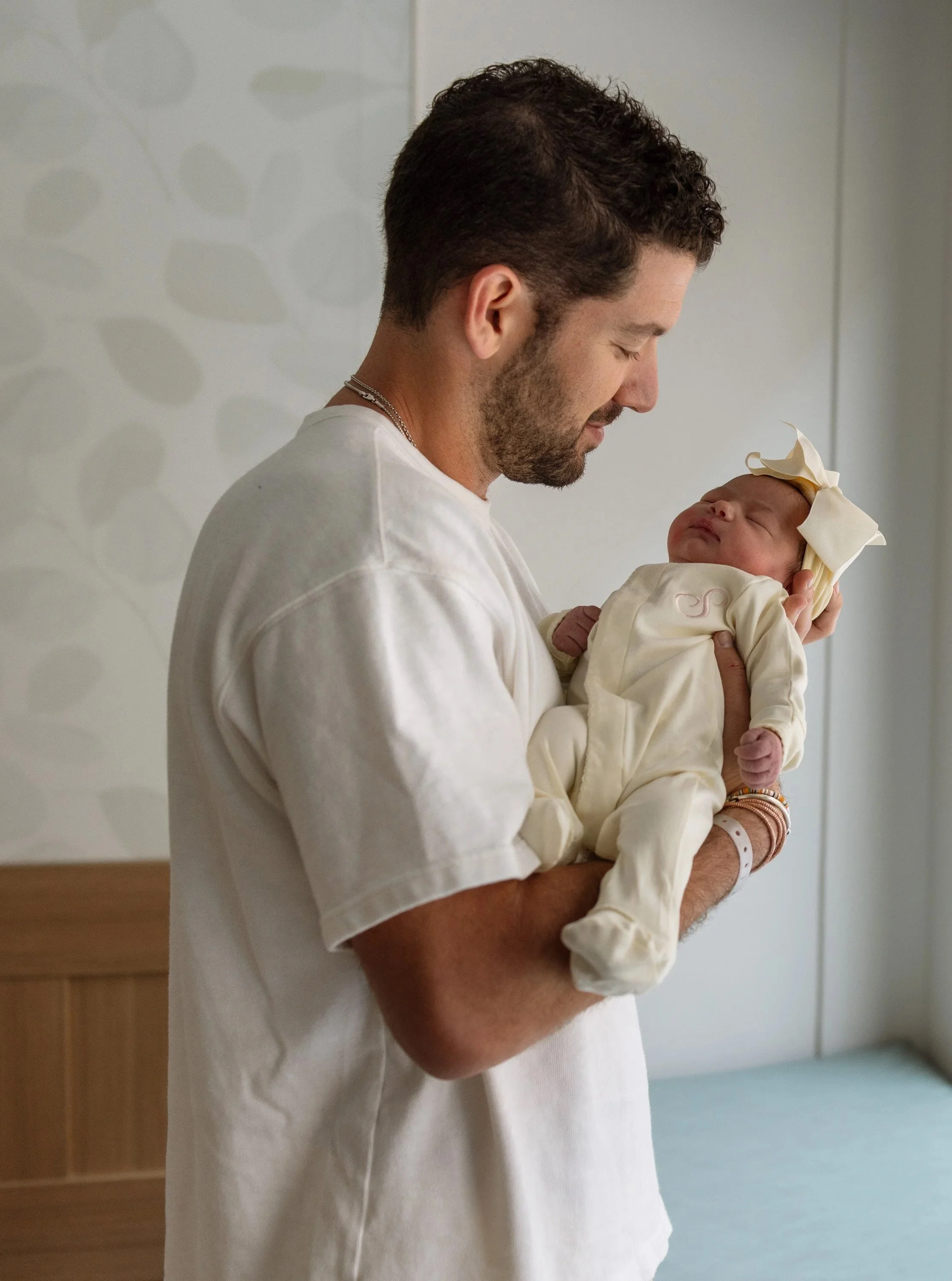 fresh 48 session in Austin Texas of a dad holding his newborn daughter looking at her and smiling. baby is wearing a butter yellow monogrammed footie outfit  paired with a matching bow headband.