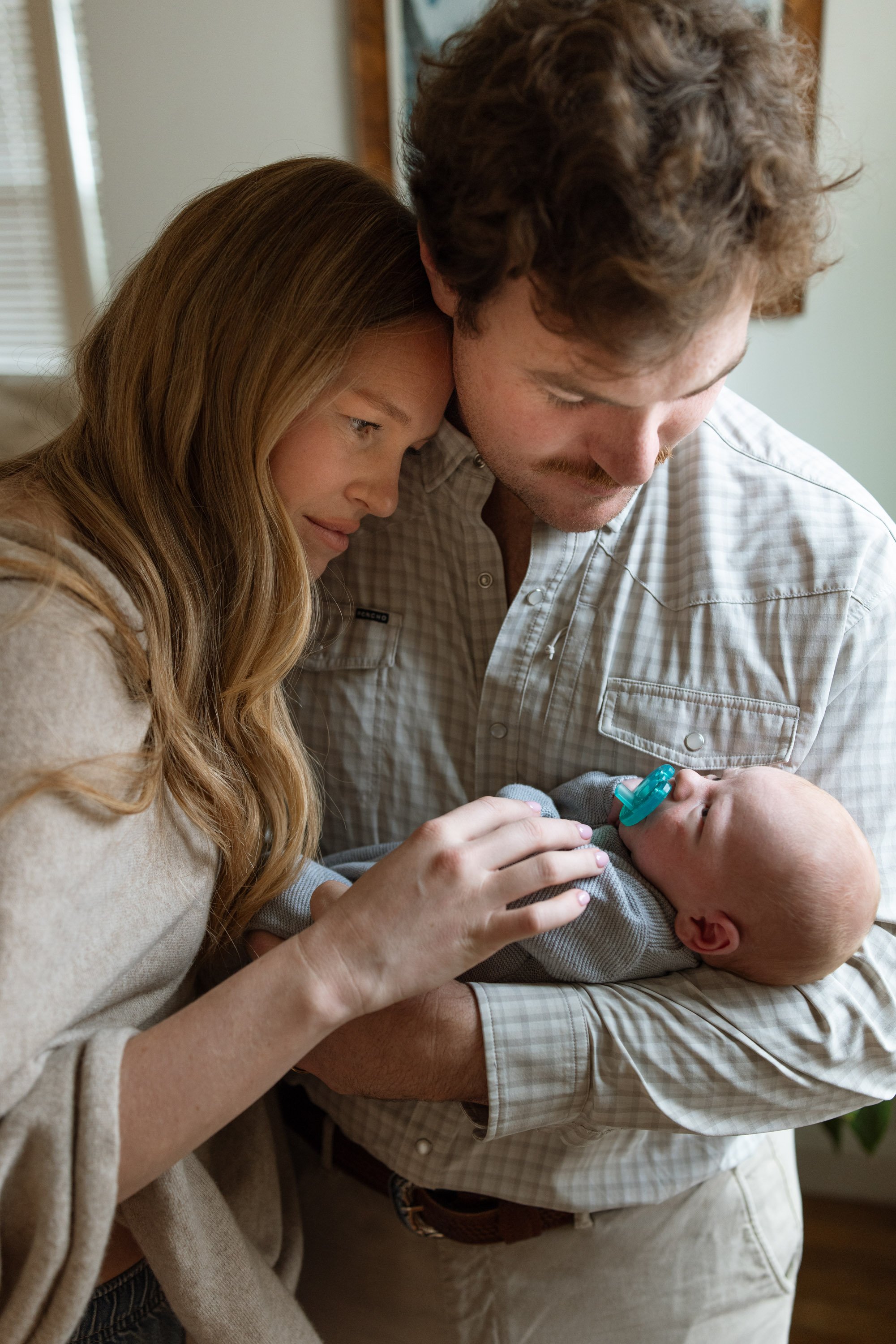 A couple standing shoulder to shoulder holding their newborn in a blue knit outfit smiling at him