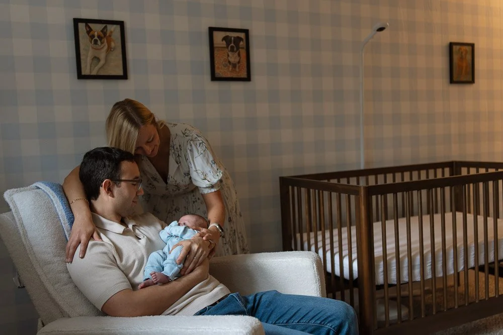 A man and a woman sitting on a couch, holding a newborn baby. The woman is leaning over, looking at the baby, while the man is seated and also looking at the baby. There is a crib in the background and framed pictures of dogs on the wall.