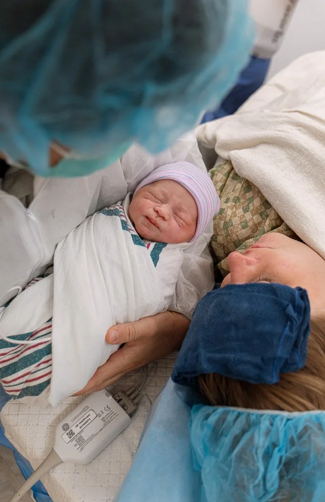 A newborn baby wrapped in a hospital blanket, wearing a pink and blue striped hat, resting in a hospital bed. An adult woman, possibly the mother, is lying beside the baby with a blue hair covering, and medical equipment is visible in the background.