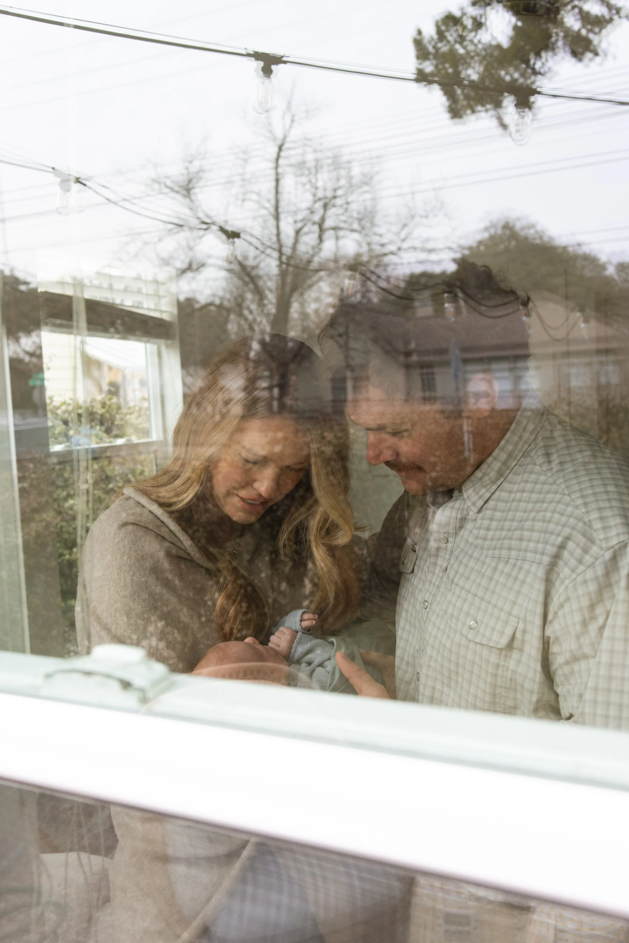 A couple holding a newborn baby inside a house, viewed through a window with reflections of trees and outdoor lights.