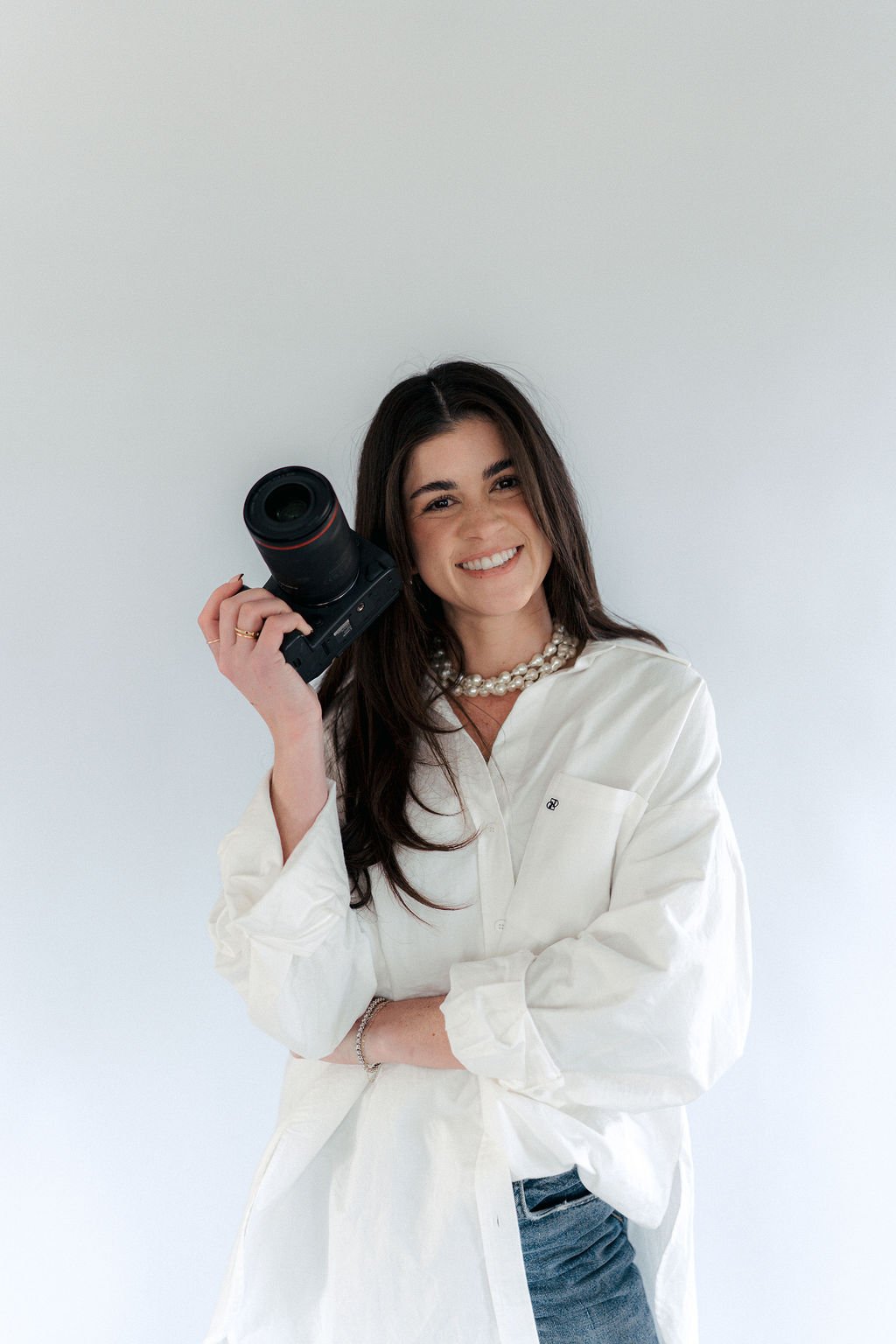 A woman smiling while holding a camera to her shoulder against a plain white background.