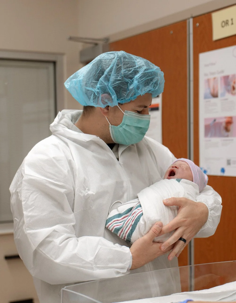 Healthcare worker in protective gear holding a newborn baby in a hospital setting.