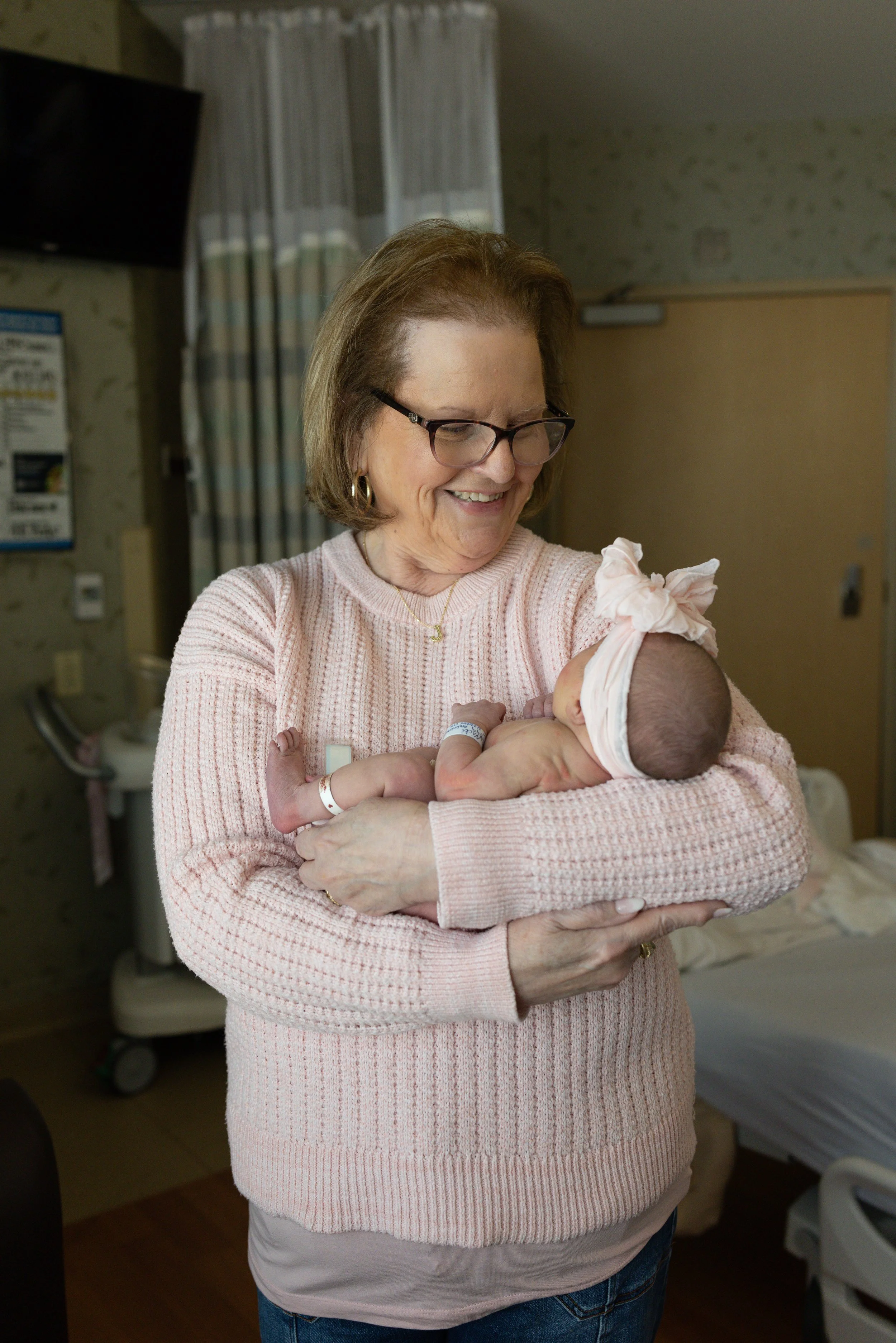 a grandma wearing a pink sweater is standing in a post-partum hospital room cradling her new granddaughter who is wearing only a pink bow headband. 