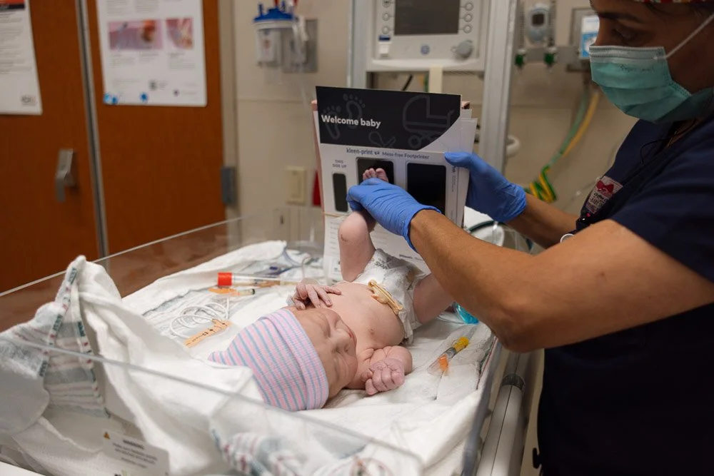 A newborn baby lying in a hospital bassinet, wearing a striped pink and blue hat, with medical tubes and wires attached. A medical professional in scrubs, gloves, and a mask is holding a brochure above the baby.