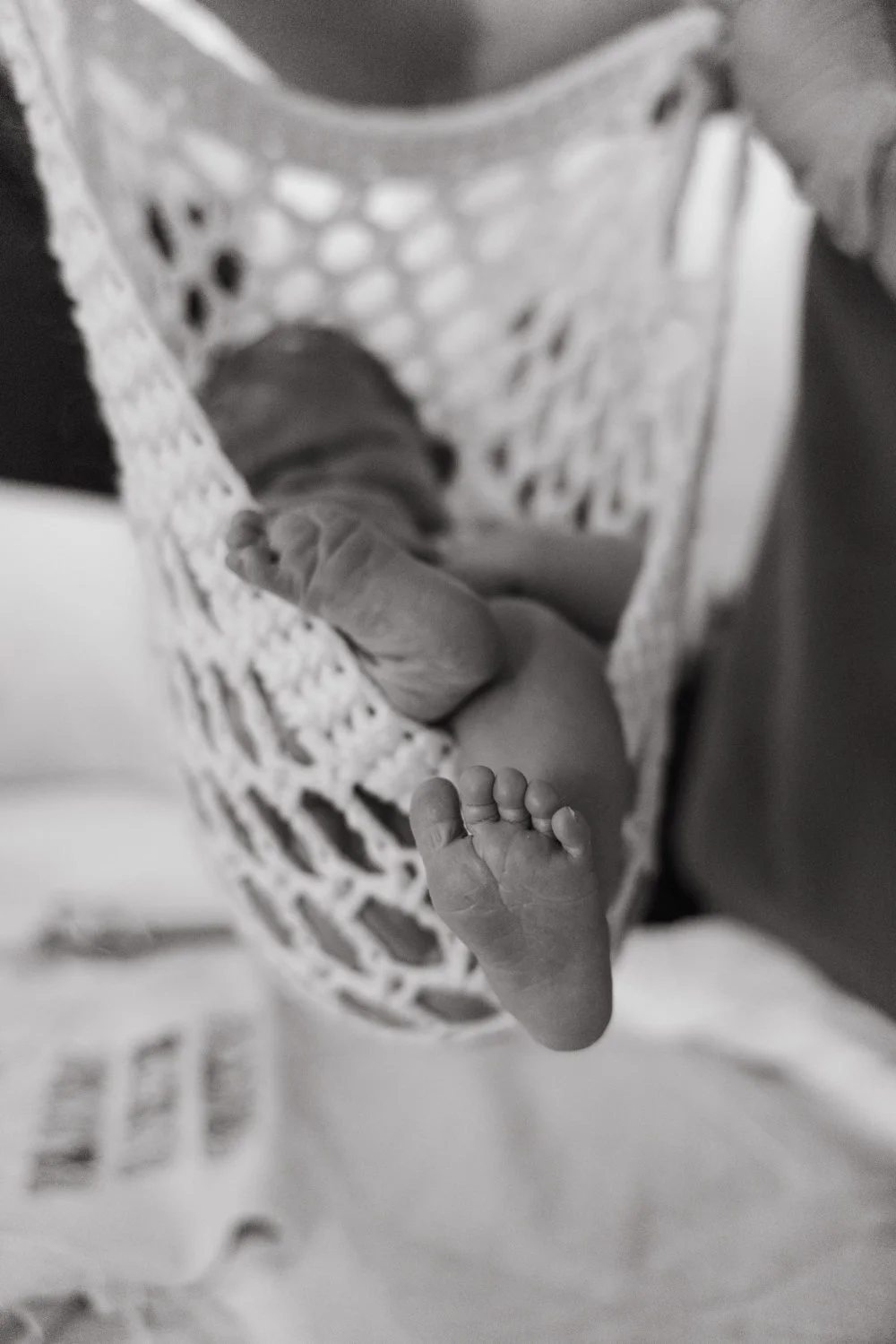 Close-up of a tiny newborn baby's foot with toes, lying on a person's arm, in a crocheted blanket and surrounded by soft fabric, in black and white.