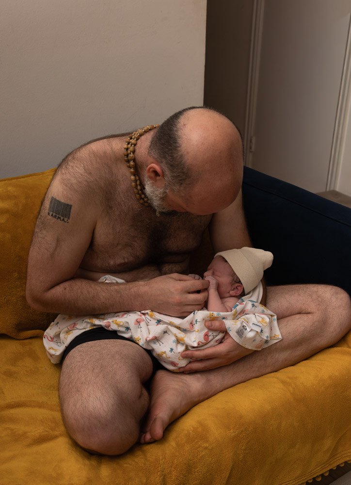 A shirtless man with a soccer tattoo on his arm, wearing a beaded necklace, lovingly holding a newborn baby wrapped in a blanket and wearing a knit cap while sitting on a yellow couch.