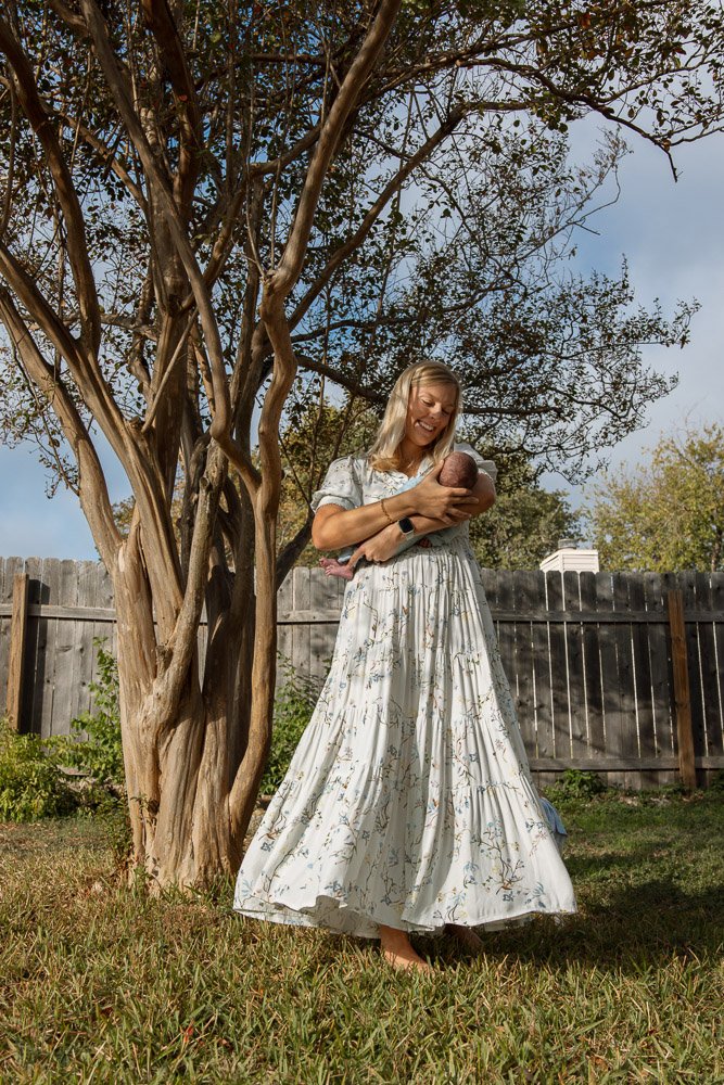 Woman in a long floral dress holding a newborn baby outdoors next to a tree in a backyard with a wooden fence.