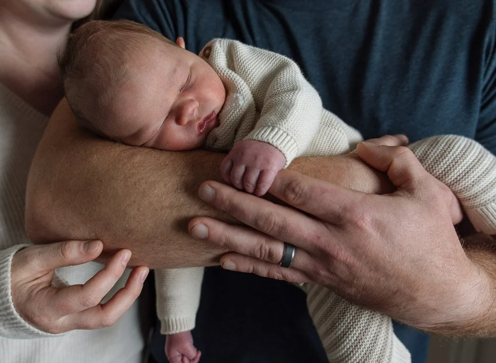 A newborn baby resting on an adult's arm with eyes closed, wearing a cream-colored sweater, in a close-up shot.