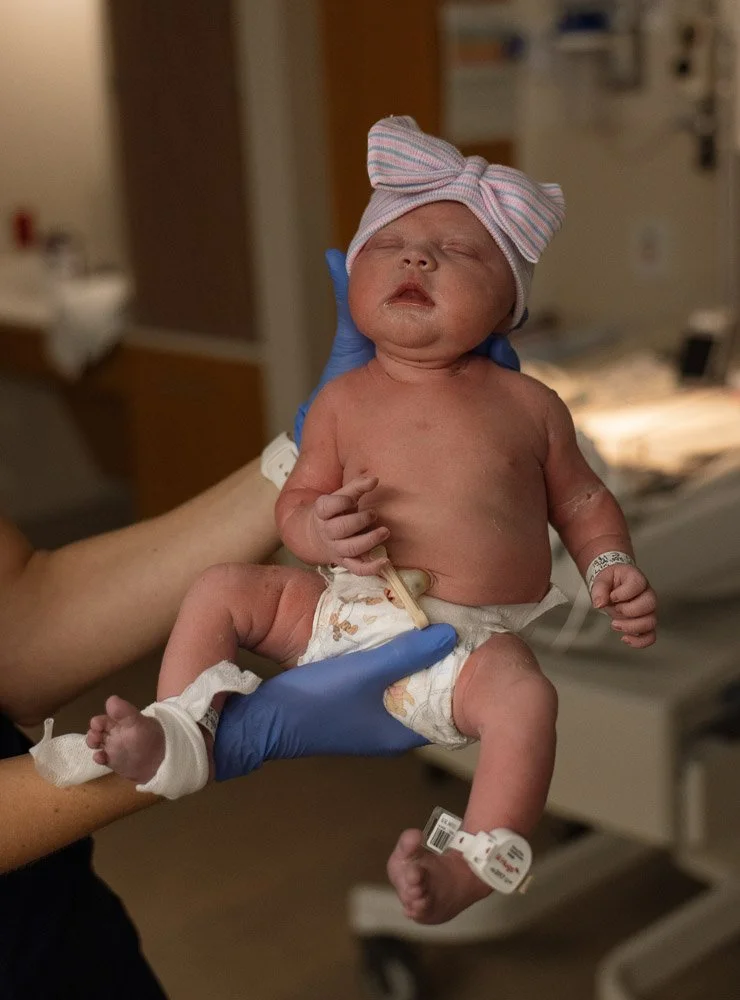A newborn baby with a pink and white striped bow hat, lying in a hospital bed while being held by a healthcare worker wearing blue gloves.