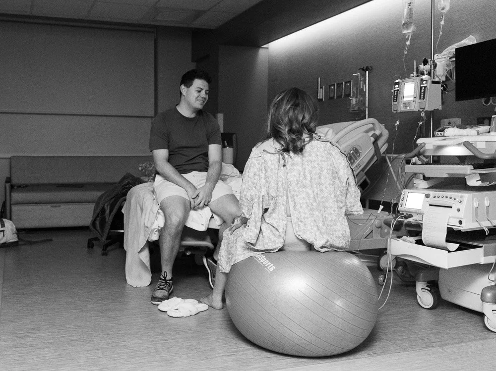 A young man is sitting on a hospital bed, smiling, as a woman, possibly a patient, sits nearby on an exercise ball in a hospital room with medical equipment and monitors.