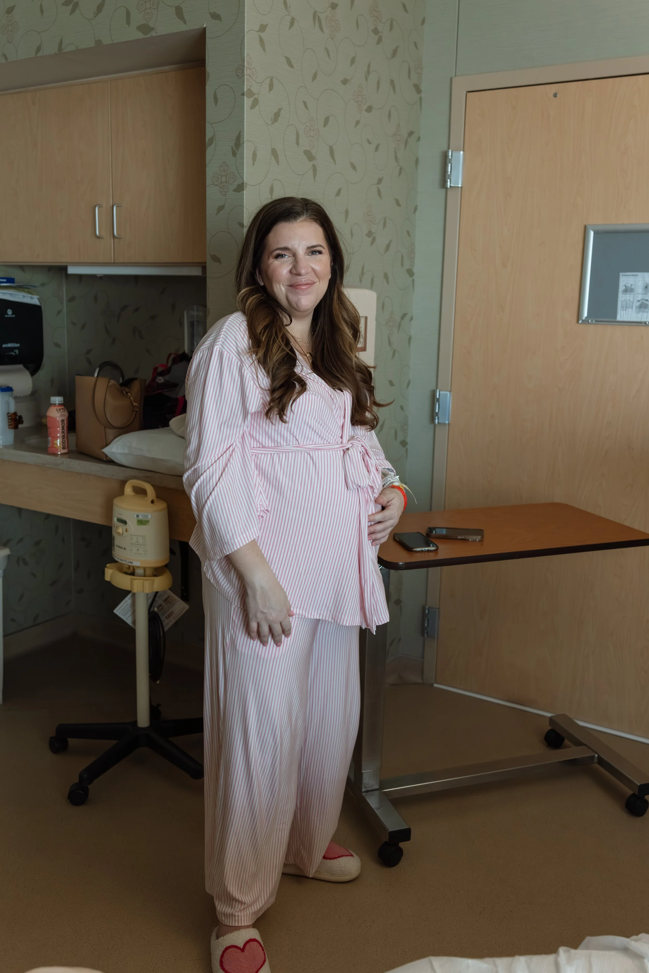 a new mom stands in a hospital room wearing coordinating pink pajamas and robe while softly smiling at the camera. there is a breast pump in the background. 
