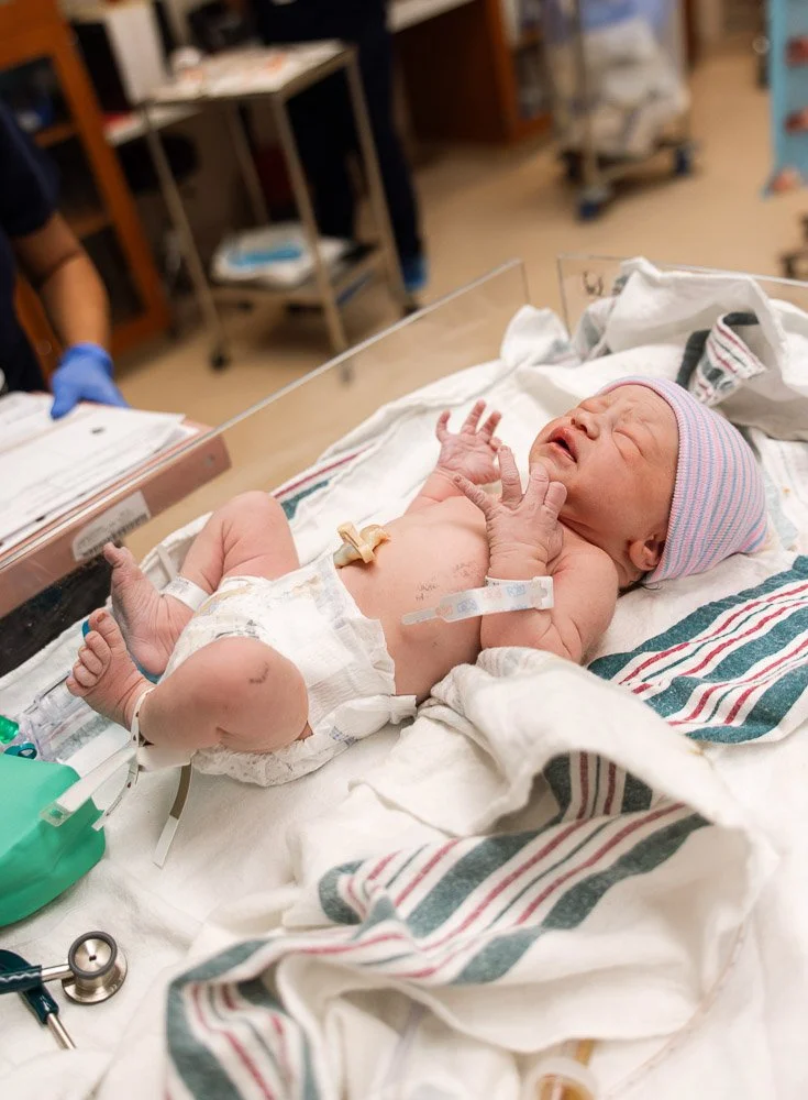 Newborn baby lying on hospital bed with medical tubes and monitors, wearing a striped hat.