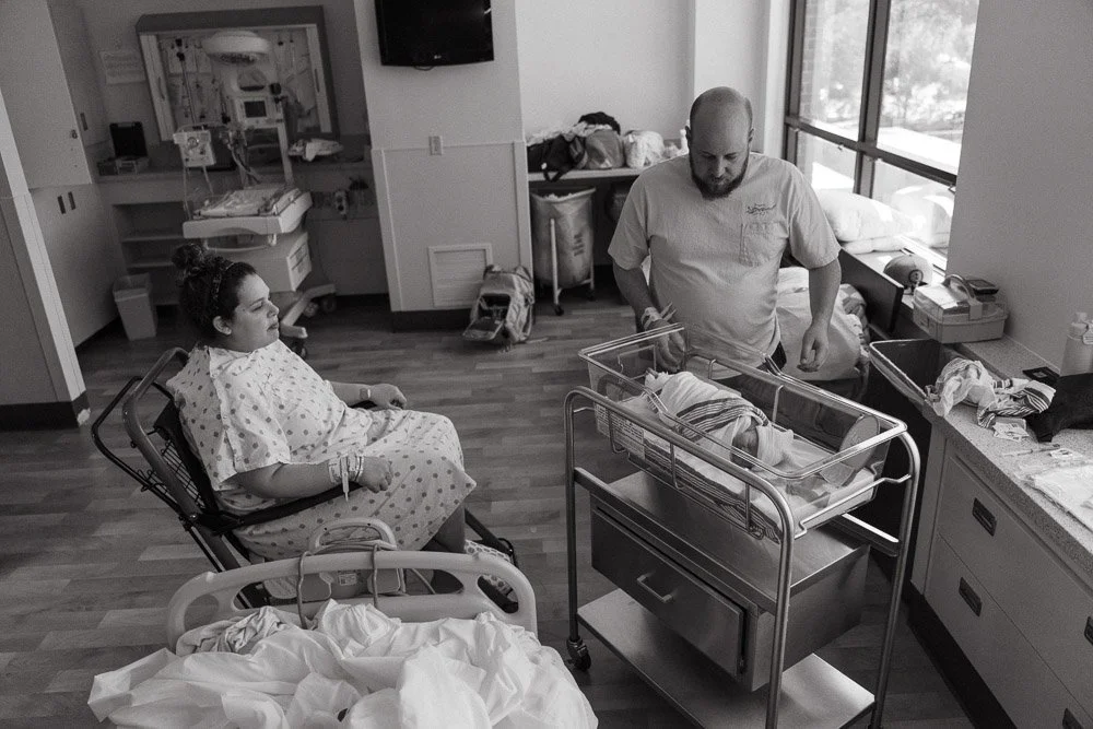 A new mother in a hospital gown sits in a wheelchair next to her newborn baby, who is in a clear bassinet. A man, likely a nurse or doctor, stands nearby tending to the baby. The room has large windows, hospital equipment, and a hospital bed.