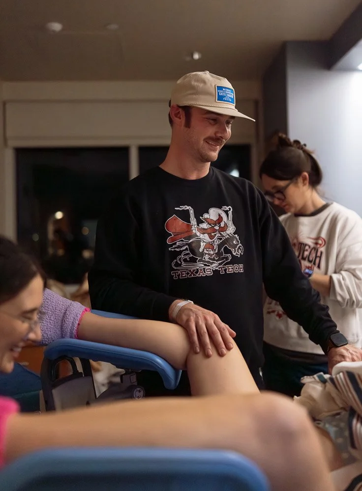 Man smiling while donating blood at a hospital, with a woman and another healthcare worker nearby.