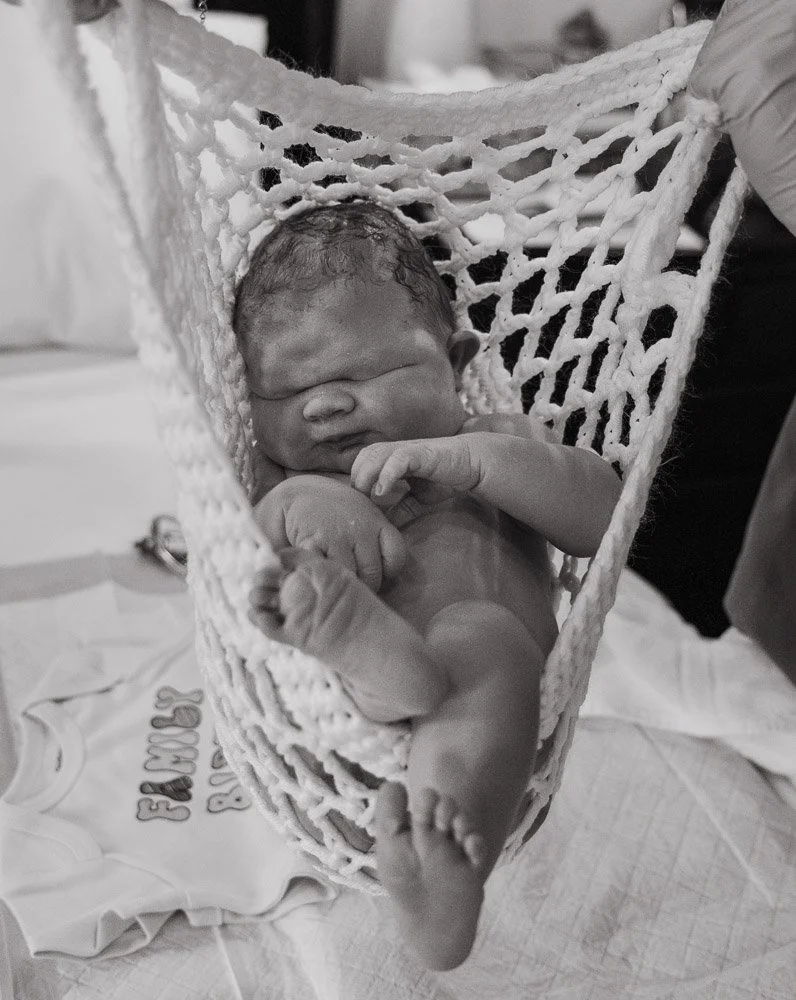 A newborn baby sleeping in a small hammock cradle, with a person gently holding and supporting the baby's head and back.