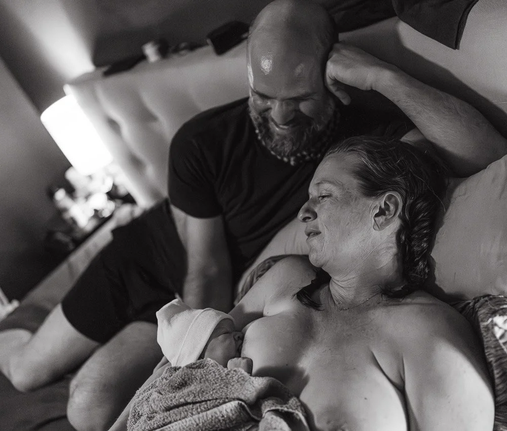 A black and white photograph of a man and woman lying on a bed, sharing a tender moment. The man is smiling and looking at the woman, who appears to be holding a newborn baby close to her chest.