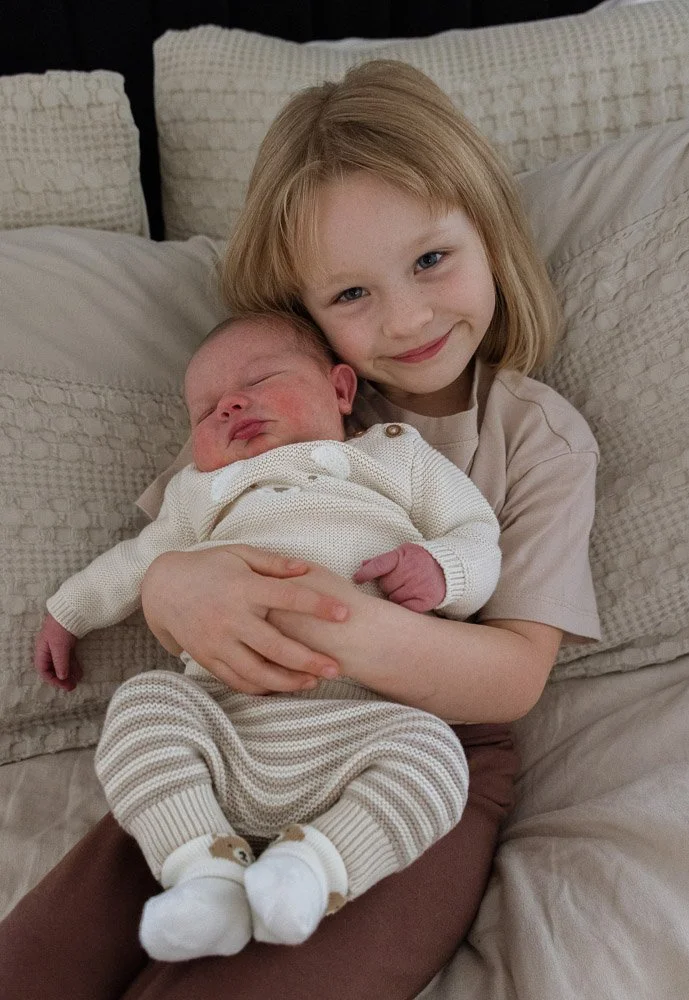A young girl holding a sleeping newborn baby, sitting on a bed with cream-colored pillows and bedding.