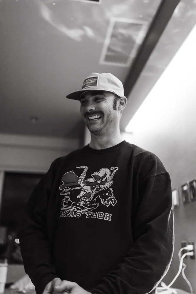 A smiling man wearing a baseball cap and a Texas Tech sweatshirt, standing indoors, with a ceiling and wall in the background.
