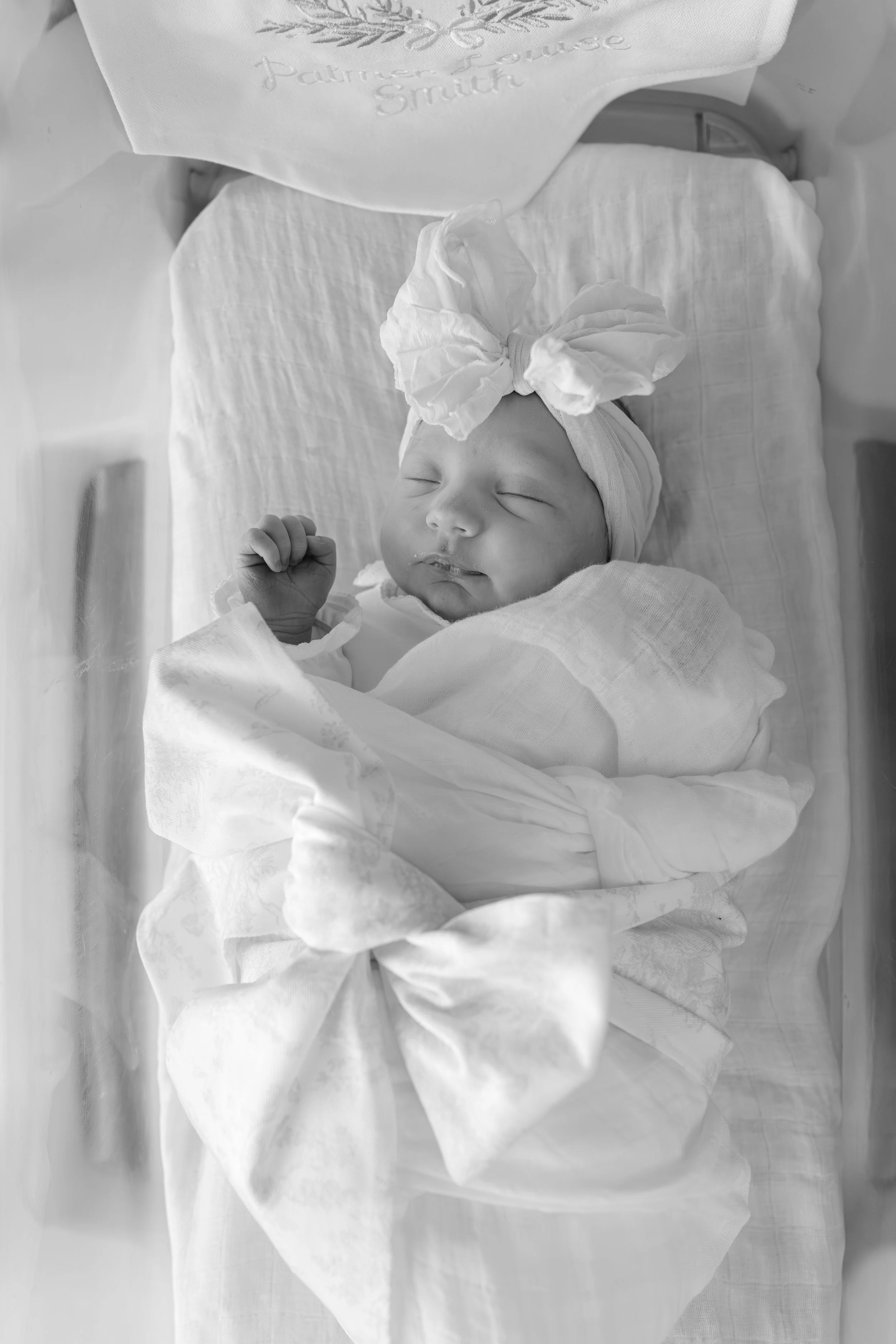 a black and white image shows a newborn baby laying in a hospital bassinet wearing a bow headband and monogrammed bow swaddle. the image is illuminated by natural light 
