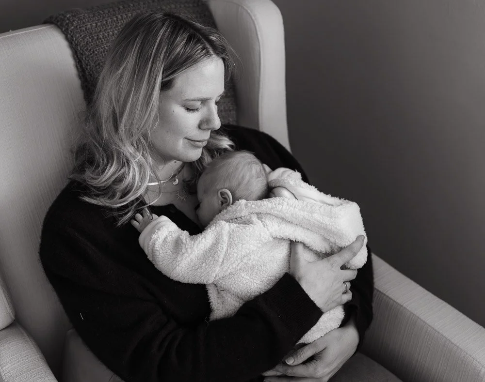 A woman with shoulder-length hair holding a baby dressed in a cozy fleece jacket, sitting on a sofa in a calm, intimate moment.